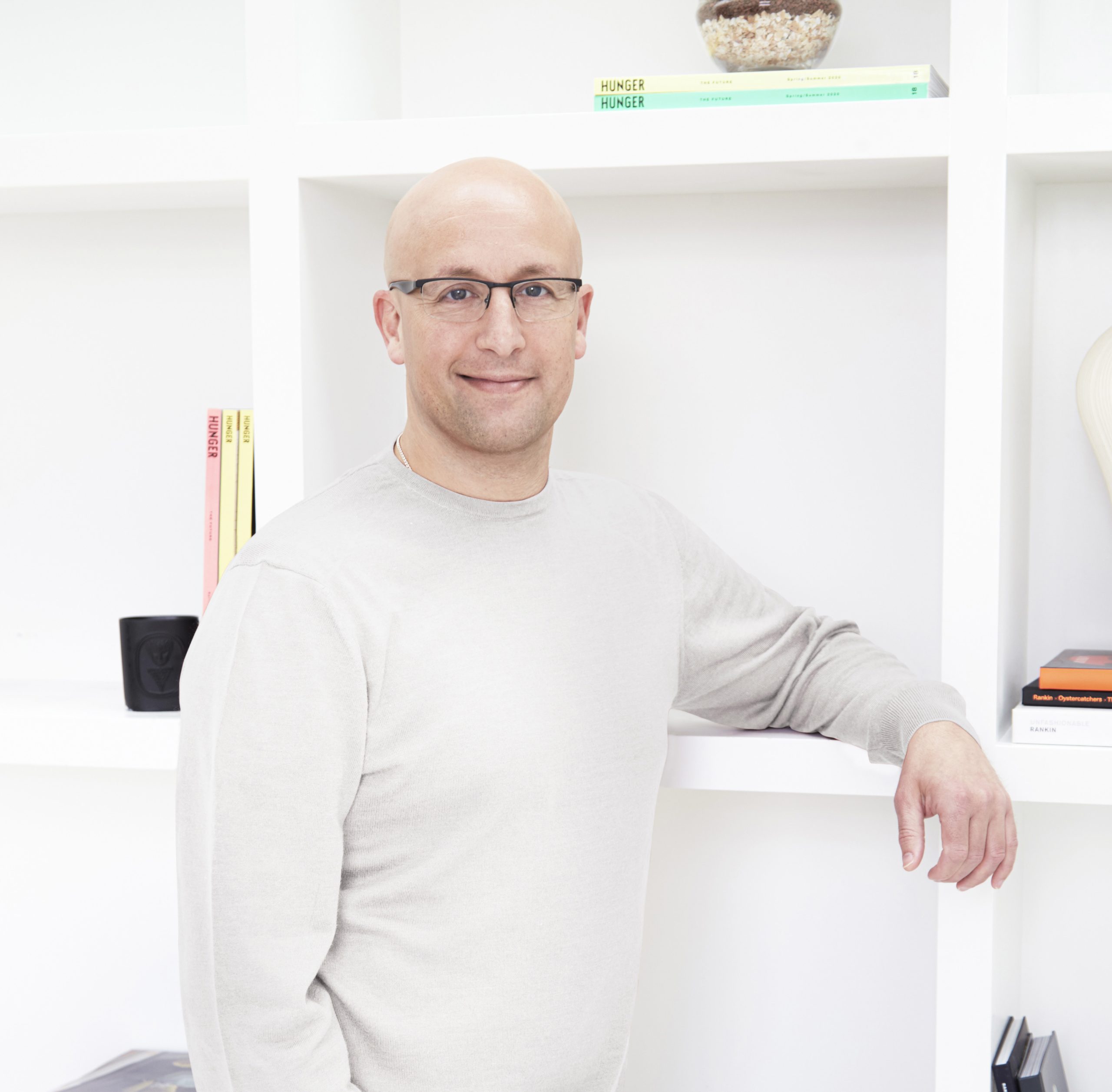 Adam Cooper wearing glasses and a off-white sweater, standing buy a white dookshelf